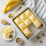 Overhead shot of a baking pan filled with pineapple banana cheesecake bars, garnished with whipped cream and walnuts, alongside ripe bananas and graham cracker crumbs.